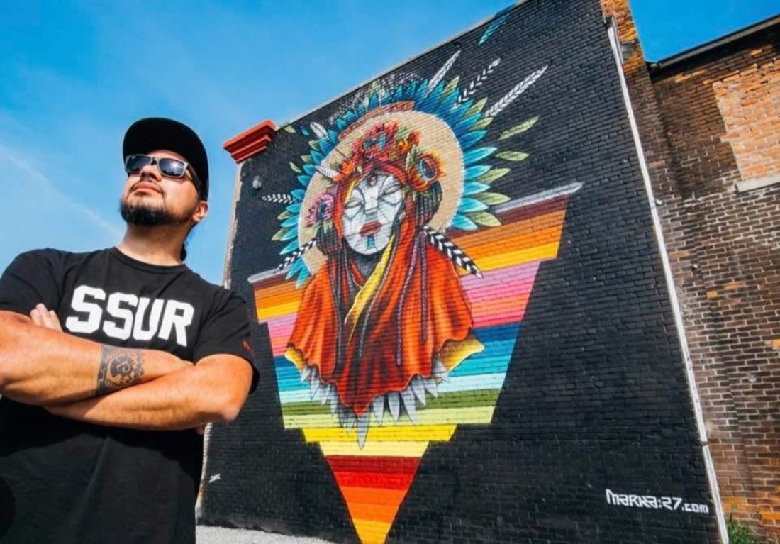 Victor MARKA27 Quiñonez standing beside a colorful Indigenous-inspired mural