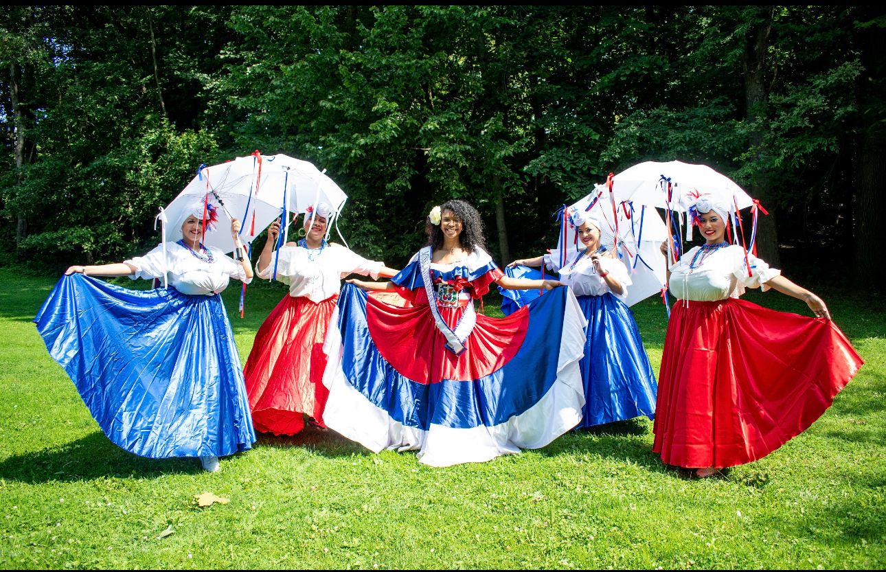 Dominican folkloric dancers pose outdoors in red, white and blue dresses.