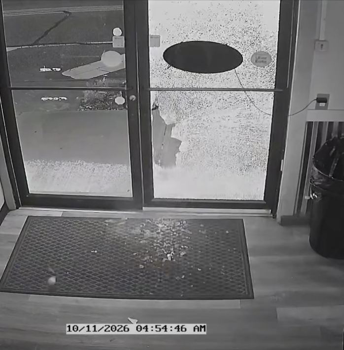 Interior surveillance view of shattered door glass and debris on a mat