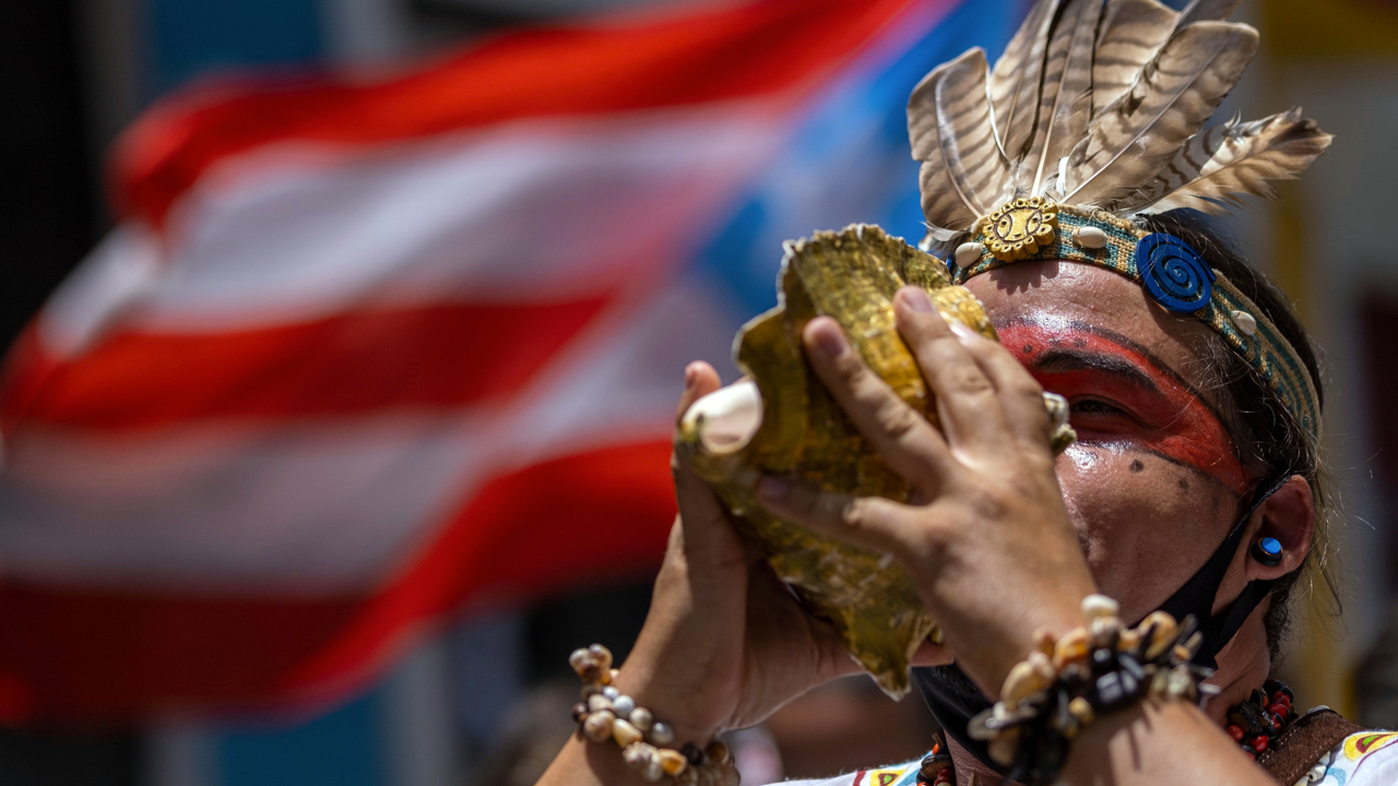 A person with feathered headwear blowing a conch shell in front of a blurred Puerto Rican flag