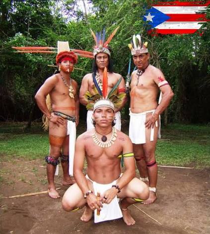 Four people outdoors wearing feathered headwear, body paint, and necklaces, with a Puerto Rican flag graphic