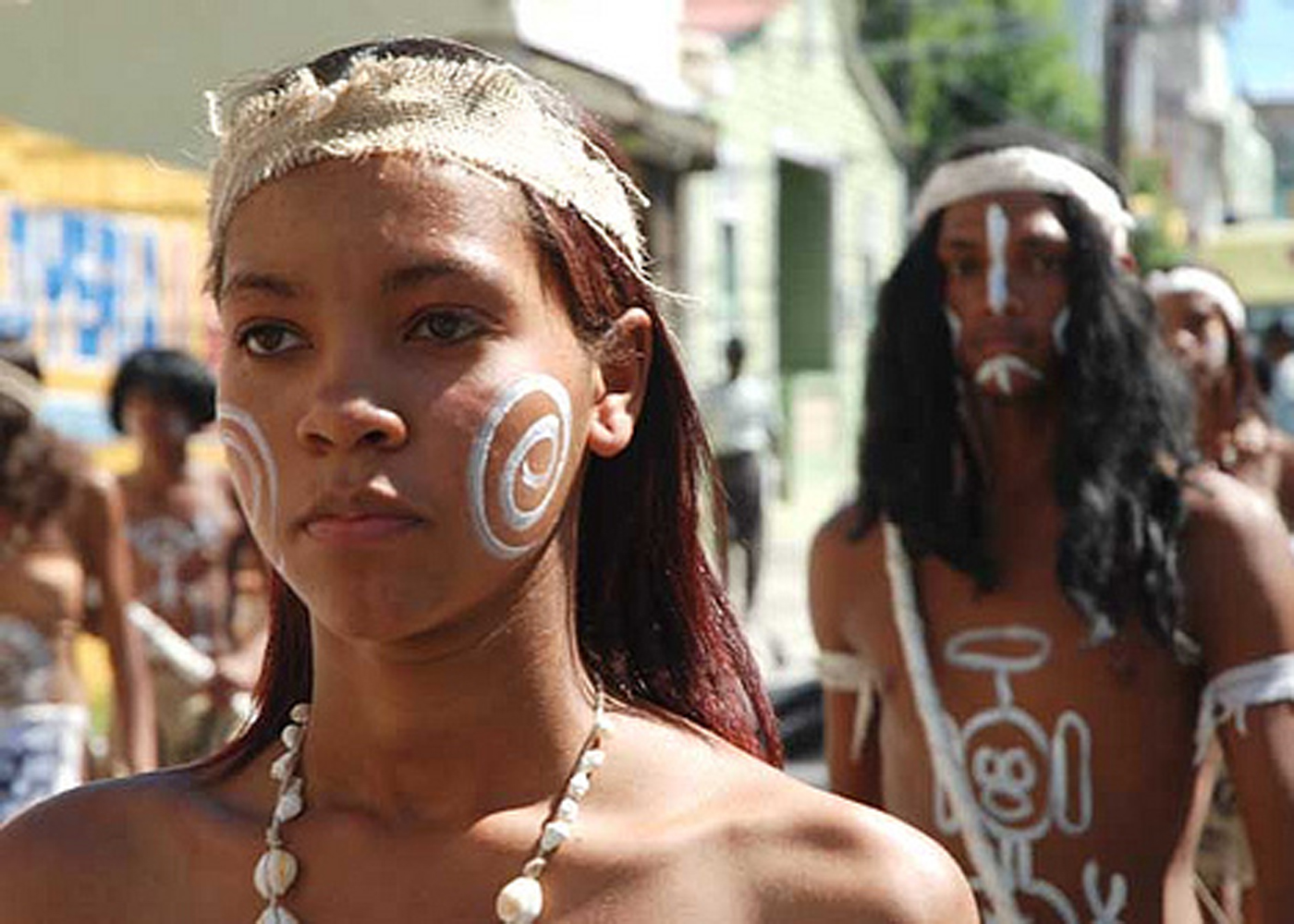 Close portrait of a young person wearing white spiral face paint and a shell necklace, with other people behind them